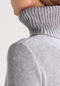 Grå stickad tröja med hög ribbad krage och textilstruktur, med tajta ärmar och en mjuk look.