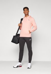 Sweatshirt à capuche rose clair avec poche avant, joggeurs gris fuselés avec logo sur le côté, et chaussures de sport blanches, tenant un sac de sport noir.