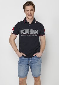Tengerészeti póló fehér és piros akcentusokkal, nagy "KRSH" logóval és "UNLIMITED BRAND 2001" felirattal. Világoskék farmer sorttal viselve.