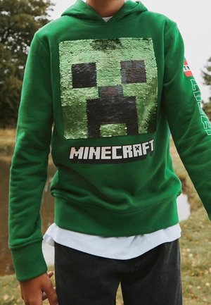 Enfant portant un sweat à capuche vert avec un motif de visage de Creeper Minecraft en sequins et un logo, debout à l'extérieur près de l'eau et des arbres.