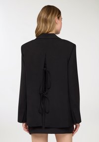 Blazer nero con vestibilità sagomata, caratterizzato da un design con spacco posteriore e nastri da annodare. Tessuto liscio e maniche lunghe. Stile semplice e minimalista.