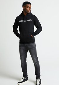 Felpa nera con tasca frontale, "ALESSANDRO" stampato in bianco. Abbinata a jeans grigi e sneakers nere con accenti bianchi.