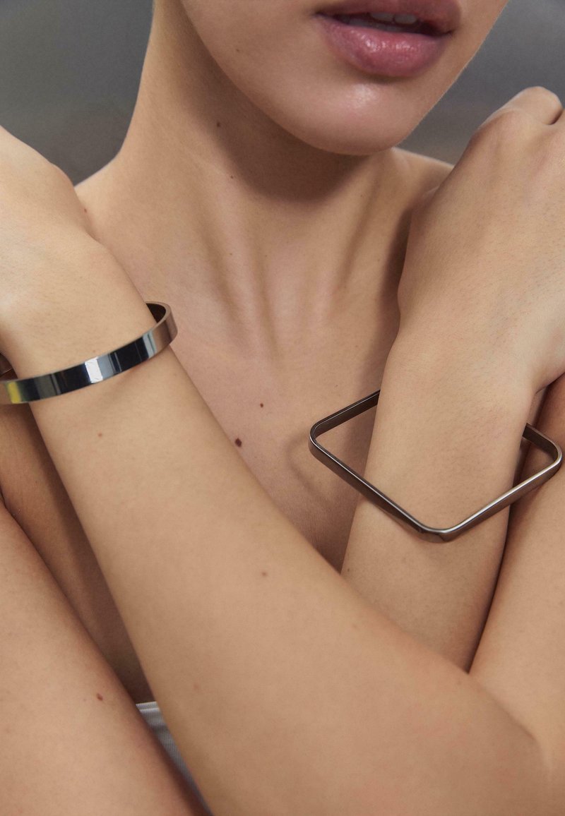 Gros plan sur des bras croisés portant un bracelet rond et un bracelet carré en argent sur une peau nue sur un fond gris.
