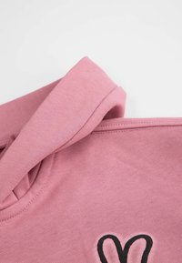 Różowy bawełniany strój z zaokrąglonym dekoltem i krótkimi rękawami. Na przodzie znajduje się czarne serce, prezentujące gładką fakturę.