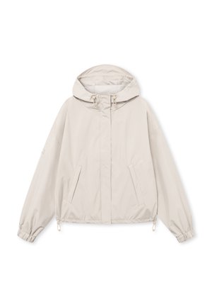 Fiatal nő világos bézs kapucnis dzsekiben, amelyen rugalmas mandzsetták vannak, és fekete nadrágot visel, egyszínű háttér előtt áll.