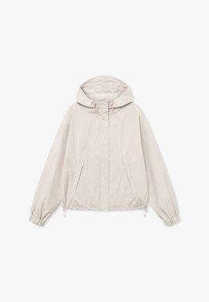 Tânără purtând o jachetă bej deschis cu glugă și manșete elastice, împreună cu o pereche de pantaloni negri, stând în picioare pe un fundal simplu.