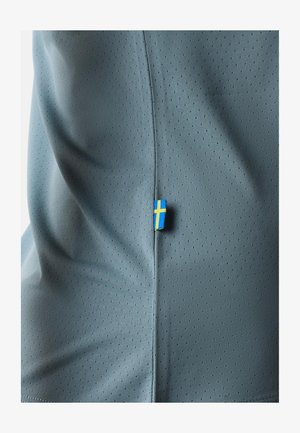 Graues Sportshirt mit strukturiertem Mesh-Design, mit einem kleinen blauen und gelben Etikett, das eine Fahne an der Seitennaht zeigt.