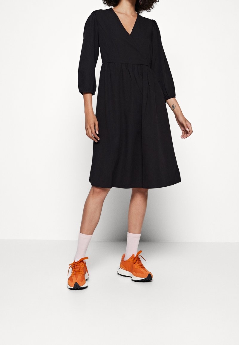 Femme portant une robe noire longueur genoux à manches trois-quarts, des chaussettes roses et des baskets orange vif, debout devant un fond blanc uni.