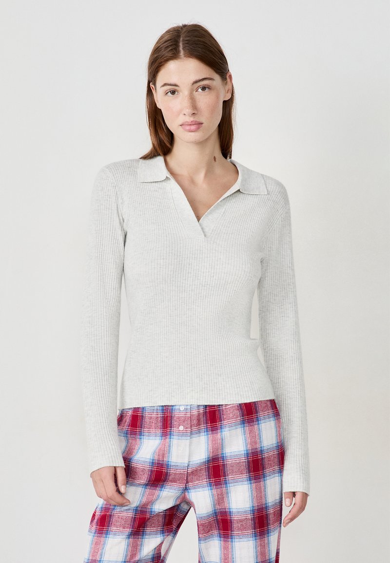 Junge Frau mit glattem braunem Haar trägt ein hellgraues, ribbeliges Langarmoberteil und rot-blau-weiße karierten Knopf-Shorts.