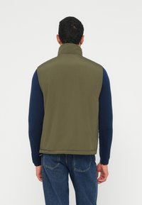 Γιλέκο χωρίς μανίκια σε ελιά, κατασκευασμένο από λείο ύφασμα, με ψηλό γιακά και ίσια γραμμή, συνδυασμένο με μπλε σκούρο πουκάμισο με μακριά μανίκια.