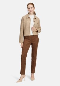 Kobieta uśmiechająca się, ubrana w beżową, teksturowaną krótką kurtkę, białą bluzkę, brązowe dopasowane spodnie oraz beżowe szpilki, stojąca na białym tle.