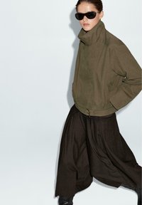 Giacca bomber verde oliva con colletto alto, abbinata a pantaloni larghi di colore marrone scuro. Il modello indossa occhiali da sole neri. Tessuto dalla consistenza liscia.