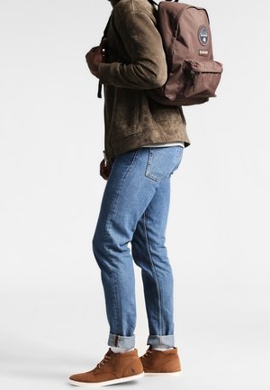 Un sac à dos en tissu brun avec un logo circulaire, porté sur une veste en daim verte, associé à un jean bleu clair et des chaussures montantes brunes.