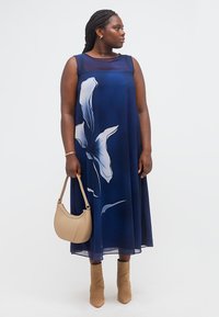 Γυναίκα με μπλε σκούρο φόρεμα χωρίς μανίκια με μεγάλο λευκό λουλουδάτο σχέδιο, κρατώντας μπεζ τσάντα, φοράει ταμπά μποτάκια στον αστράγαλο, κοιτάζει προς τα δεξιά.