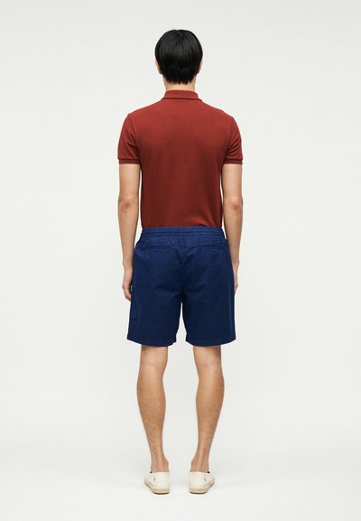 Άνδρας που στέκεται με την πλάτη γυρισμένη φοράει πορτοκαλί πόλο, μπλε σορτς και λευκά παπούτσια slip-on, πάνω σε λευκό απλό φόντο.