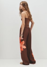Giovane donna con capelli lunghi che indossa una canotta marrone e pantaloni larghi marroni con grandi stampe di fiori arancioni, in piedi di profilo.