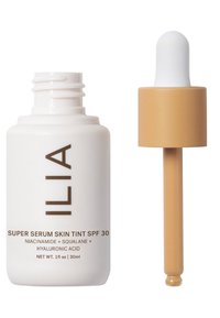 ILIA Super Serum Skin Tint SPF 30 i en vit flaska med brunt text, tillsammans med ett tan dropperlock. 30 ml storlek, med en jämn textur.