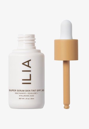 ILIA Super Serum Skin Tint SPF 30 in een witte fles met bruine tekst, gecombineerd met een tan kleurige pipetdop. 30 ml grootte, met een gladde textuur.