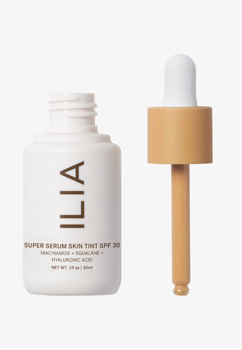 Super Serum Skin Tint SPF 30 od ILIA w białej butelce z brązowym napisem, z beżową nakrętką w kroplomierzu. Pojemność 30 ml, o gładkiej teksturze.