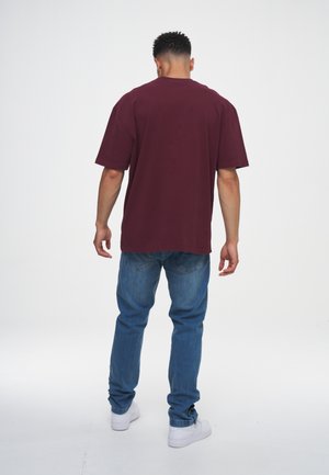 Mężczyzna stojący tyłem, ubrany w bordową, oversize'ową koszulkę, niebieskie dżinsy i białe tenisówki na jednolitym białym tle.