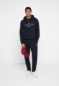 Granatowa bluza z kapturem z białym logo, dopasowane granatowe spodnie dresowe oraz czerwona czapka z daszkiem trzymana w ręku. Tkanina wydaje się miękka, a krój luźny.