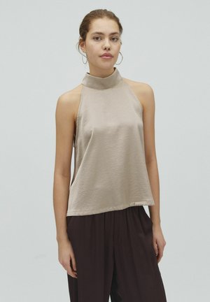 Žena v svetlo béžovom saténovom bezrukávovom halter topu a voľných tmavohnedých nohaviciach, stojaca pred hladkým svetlosivým pozadím.