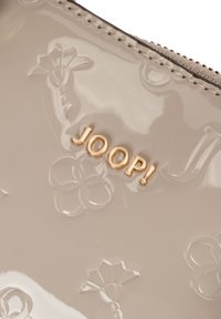 Materiale beige lucido con motivi floreali in rilievo e lettere dorate che formano la scritta "JOOP!" vicino a un bordo con zip cucito.