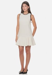 Abito bianco con orlo a campana, tessuto strutturato e rifinitura nera sul collo. Modella che indossa scarpe slingback nere.