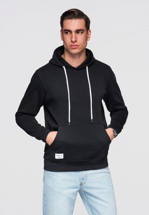 Homme portant un sweat à capuche noir avec des cordons blancs et un jean bleu clair, mains dans les poches, regardant vers sa gauche sur un fond uni.