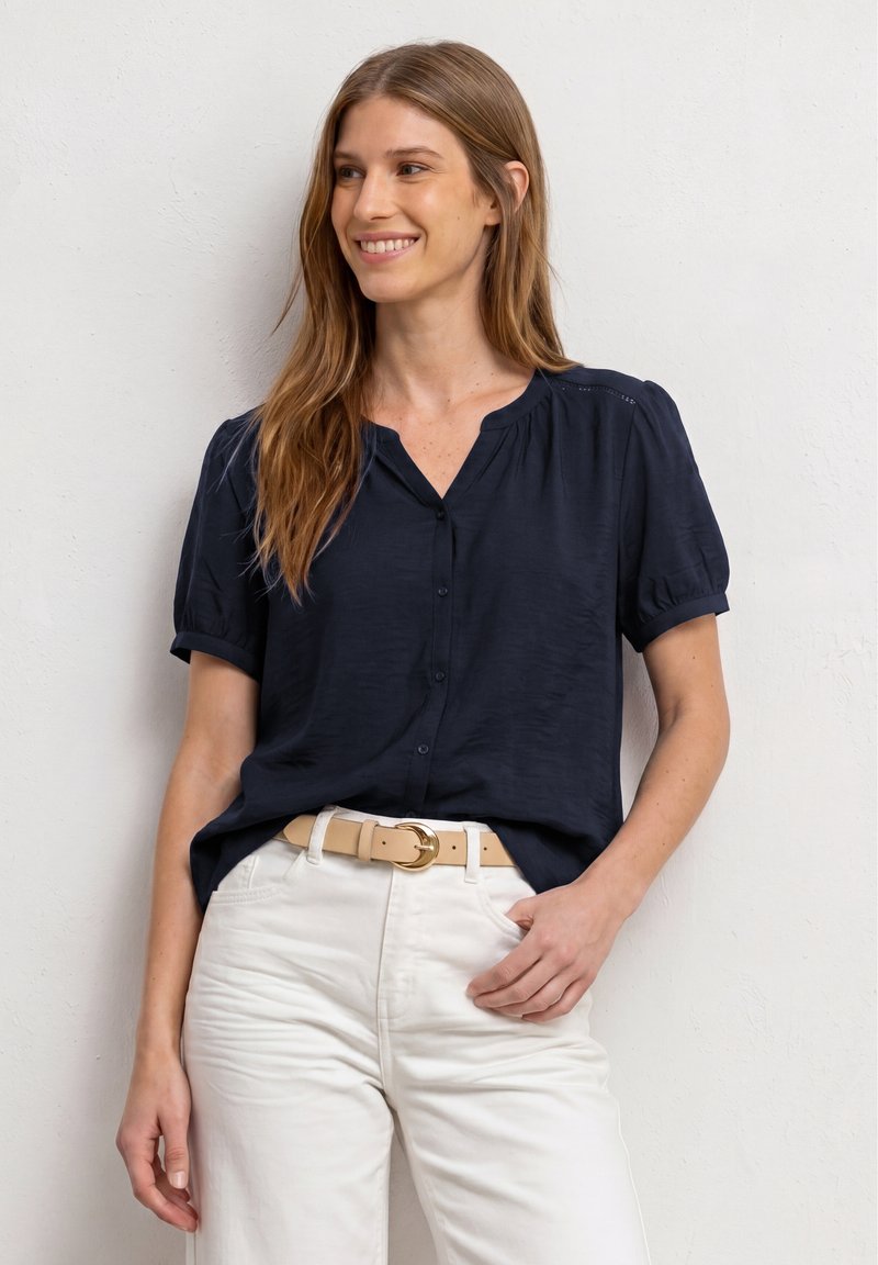 Femme souriante aux longs cheveux châtain clair portant un chemisier bleu marine à manches courtes boutonné, un pantalon blanc et une ceinture beige avec une boucle dorée.