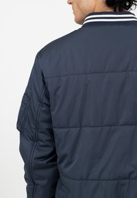 Marineblauwe gewatteerde jas met horizontaal gestikt ontwerp, geribbelde kraag met witte strepen en een zijzakdetail.