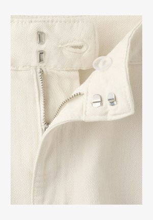 Pojas bijelih traperica s djelomično otvorenim srebrnim patentnim zatvaračem i dvostrukim metalnim kukicama i štapnim kopčama na zatvaraču.