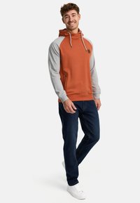 Oranje en grijze hoodie met een trekkoord, zijzakken en een ronde logo. Draagt met donkerblauwe jeans en witte sneakers.