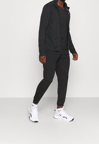 Zwarte ritssportjack en bijpassende broek van lichtgewicht materiaal, gecombineerd met witte sportschoenen met zwarte accenten en logo.