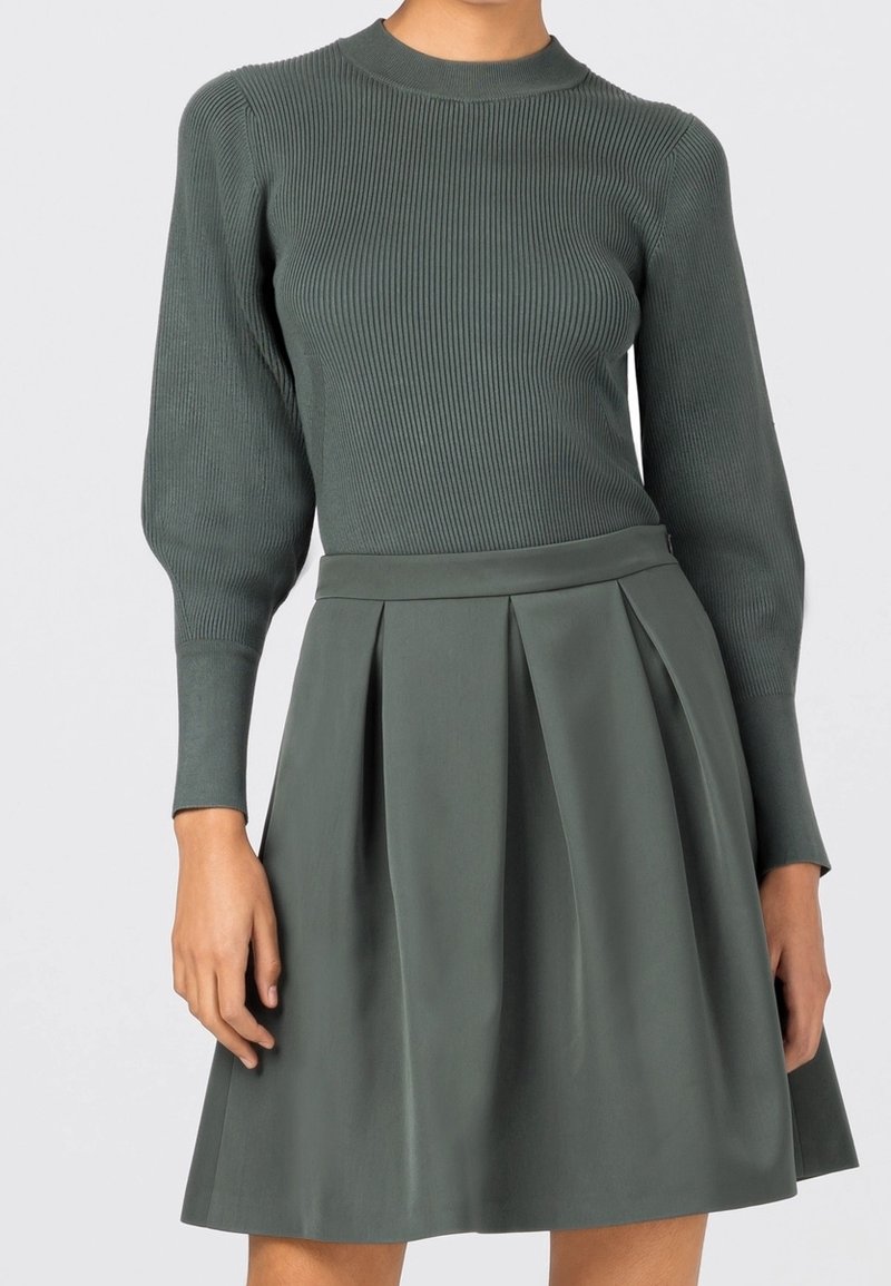 Femme portant un pull vert côtelé à manches longues associé à une jupe verte plissée taille haute sur un fond uni.