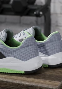 Paire de chaussures de sport grises et vertes placées sur un banc en bois avec un support d'haltères flou en arrière-plan.