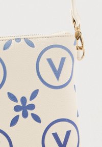 Nærbillede af beige tekstureret håndtaske med blå cirkulære "V"-logoer og blomsterprint, med en guldfarvet lås og justerbar rem.