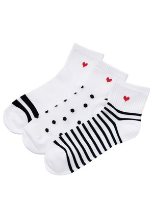 Trzy białe skarpetki do kostki z czarnymi paskami, czarnymi kropkami i czarnymi wzorami linii, z których każda ma małe czerwone serce przy ściągaczu.