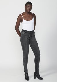 Črne ozke kavbojke z nahrbtnim detajlom in okrašenimi žepi, v kombinaciji z belo prilegajočo majico brez rokavov in črnimi gležnjarji.