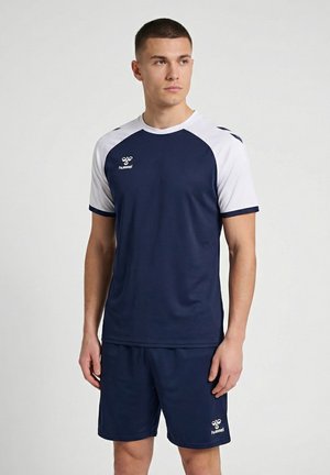 Jonge man in marineblauw en wit sportkleding, staand met ontspannen armen, kijkt iets opzij tegen een effen achtergrond.