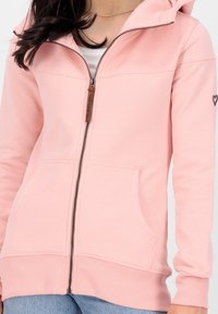Donna che indossa una felpa leggera rosa con zip, tasche frontali, zip parzialmente aperta, piccolo cuore nero sulla manica sinistra e jeans azzurri.