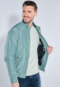 Hellgrüne Jacke mit einem Reißverschluss vorne, zwei Seitentaschen und einem schwarzen gemusterten Innenfutter. Der Stoff sieht glatt und leicht aus.