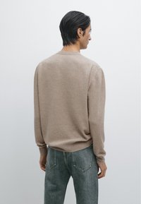 Beżowy sweter z długimi rękawami, okrągłym dekoltem, miękką fakturą, noszony na szarych dżinsach, widoczny z tyłu na jednolitym tle.