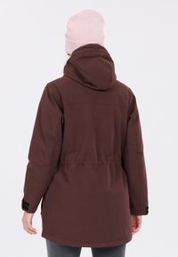 Bordeaux parka-jack met capuchon, voorzien van een getailleerde taille, zijzakken en knoopmanchetten. Draagt een lichtroze gebreide beanie.
