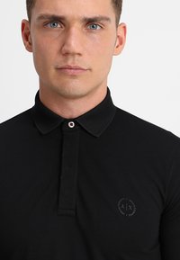 Polo nero con colletto testurizzato, chiusura con bottoni e logo sul lato sinistro del petto. Il tessuto appare liscio e aderente.