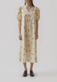 Femme portant une robe midi crème avec des manches ballon et un motif floral jaune, associée à des chaussures noires à enfiler, debout sur un fond uni.
