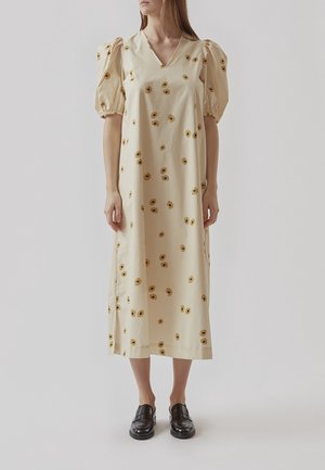 Mujer vestida con un vestido midi crema con mangas abullonadas y un patrón floral amarillo, combinado con zapatos negros tipo slip-on, de pie sobre un fondo liso.
