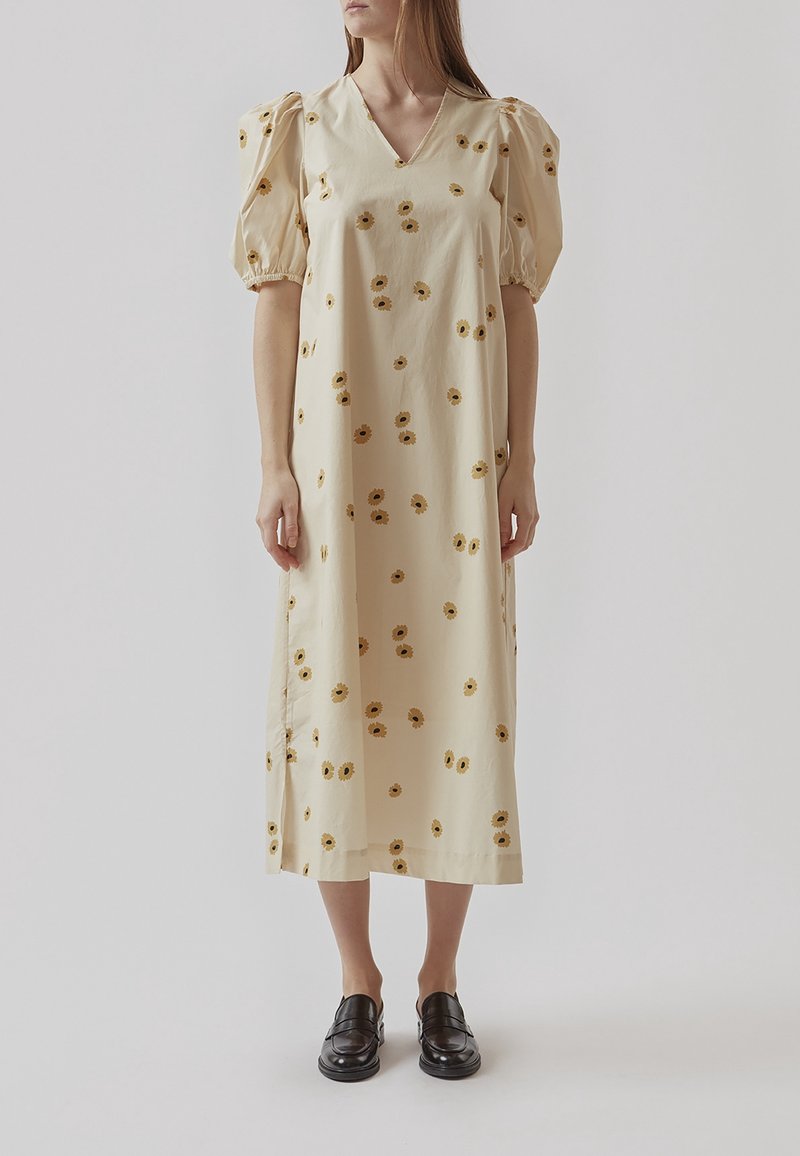 Femme portant une robe midi crème avec des manches ballon et un motif floral jaune, associée à des chaussures noires à enfiler, debout sur un fond uni.