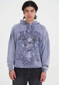 Joven con cabello negro de longitud media que lleva una sudadera morada con cruces, un corazón con "Los Ángeles" y diseños de tatuajes de rosas.