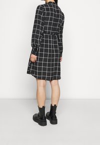 Černé šaty s dlouhým rukávem a bílým kostkovaným vzorem, se staženým pasem, s kulatým límcem, doplněné černými kotníkovými botami.